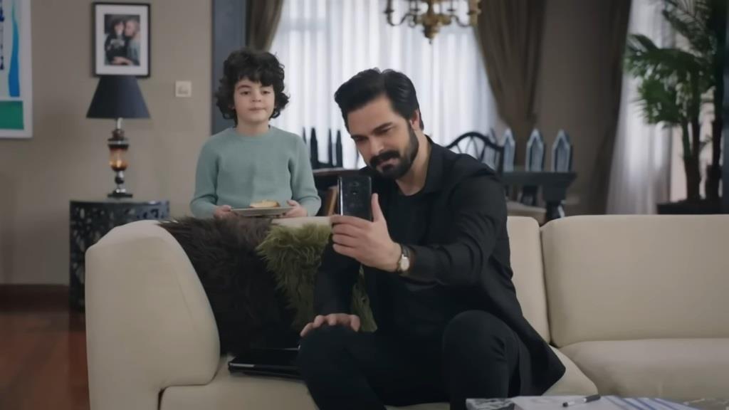 Yaman siedzi na kanapie i trzyma wyciągnięty przed siebie telefon, jakby rozmawiał z kimś przez wideorozmowę. Z tyłu, za kanapą, znajduje się Yusuf.