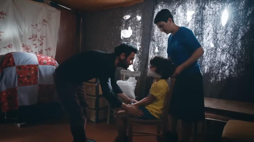 Yusuf jest związany i zakneblowany. Porywacz pochyla się nad nim i mówi coś do niego.