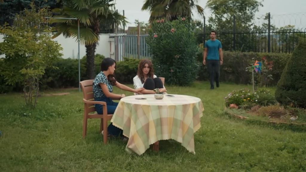 Yasemin rozmawia z Duygu, siedząc przy stole na zewnątrz. W ogrodzie stoi Ali i obserwuje je.