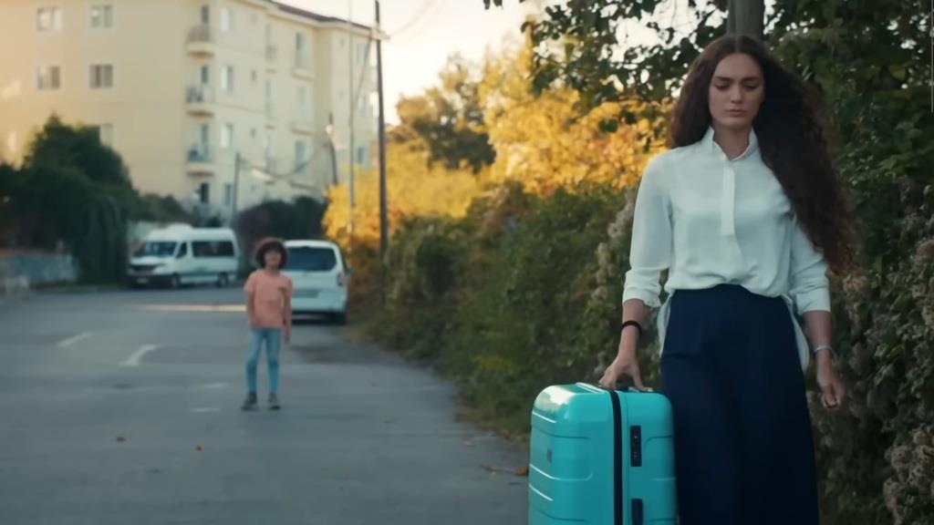 Nana idzie drogą, ciągnąc walizkę. Na drugim planie znajduje się Yusuf, który wydaje się do niej wołać.