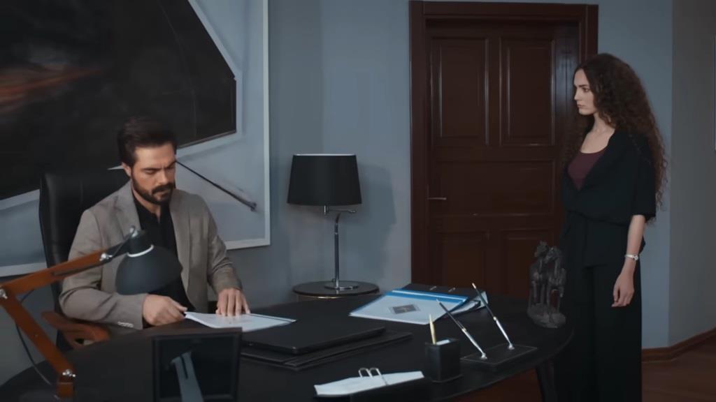 Scena w gabinecie. Yaman siedzi przy biurku. Obok stoi Nana.
