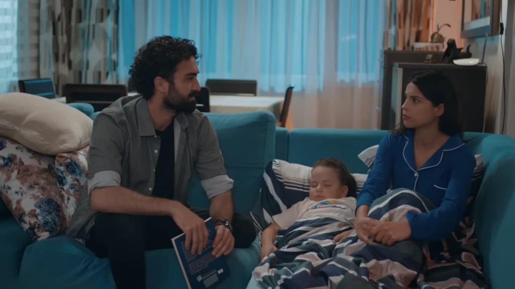Ferit trzyma książkę w rękach i patrzy na Ayse z uśmiechem. Między nimi, na kanapie, zasnęła Doga.