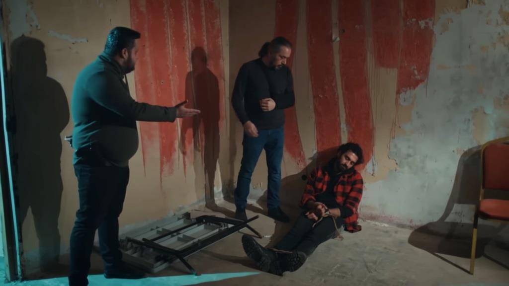 Ferit siedzi pod ścianą ze związanymi rękami i nogami. Wydaje się, że jest nieprzytomny. W pomieszczeniu znajdują się także dwaj jego porywacze.
