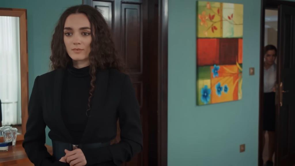 Nana stoi w pokoju, nie widząc Aynur, która obserwuje ją przez uchylone drzwi.