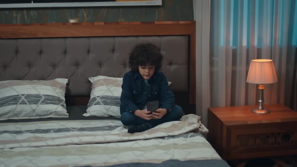 Yusuf siedzi po turecku na łóżku, trzymając w rękach telefon Cengera.