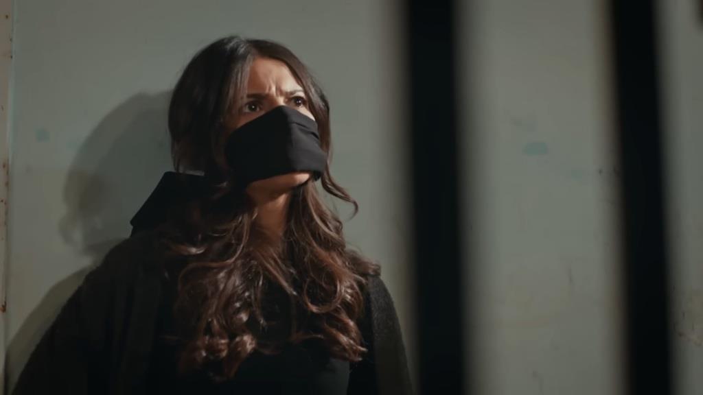 Ayse siedzi pod surową ścianą, z zakneblowanymi ustami.