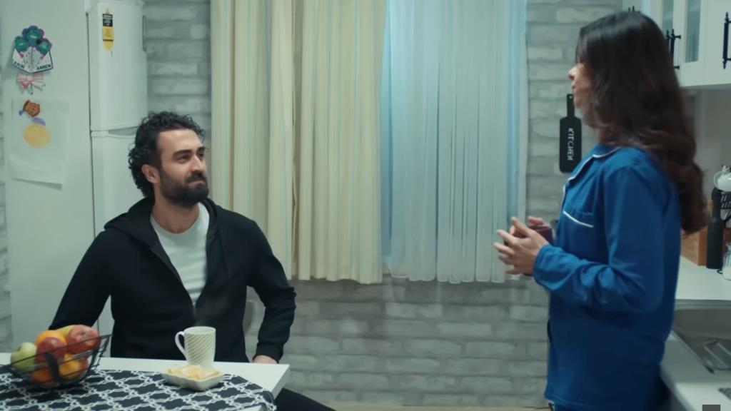 Ferit i Ayse rozmawiają w kuchni. On siedzi przy stole, ona stoi, opierając się o blat.