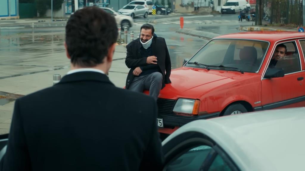 Idris w gorsecie ortopedycznym siedzi na masce czerwonego sedana i patrzy z uśmiechem na Nedima.