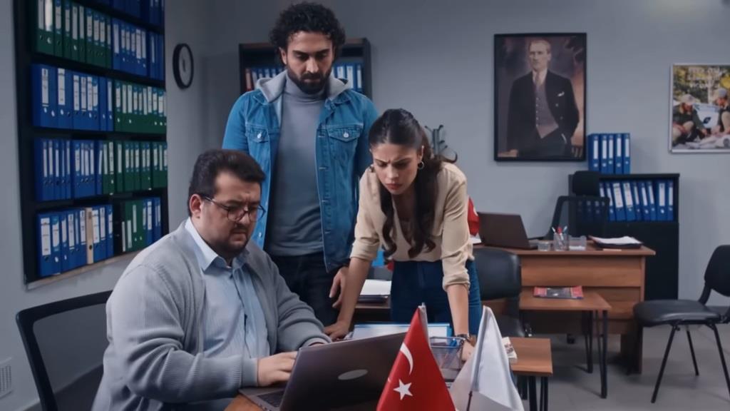 Ibrahim siedzi przy biurku i stuka w klawiaturę laptopa. Obok stoją Ayse i Ferit, także wpatrując się w ekran komputera.