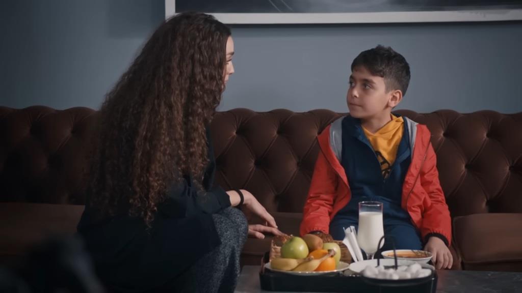 Nana siedzi obok chłopca. Na stoliku przed nim stoi szklanka z mlekiem, owoce oraz talerz zupy.