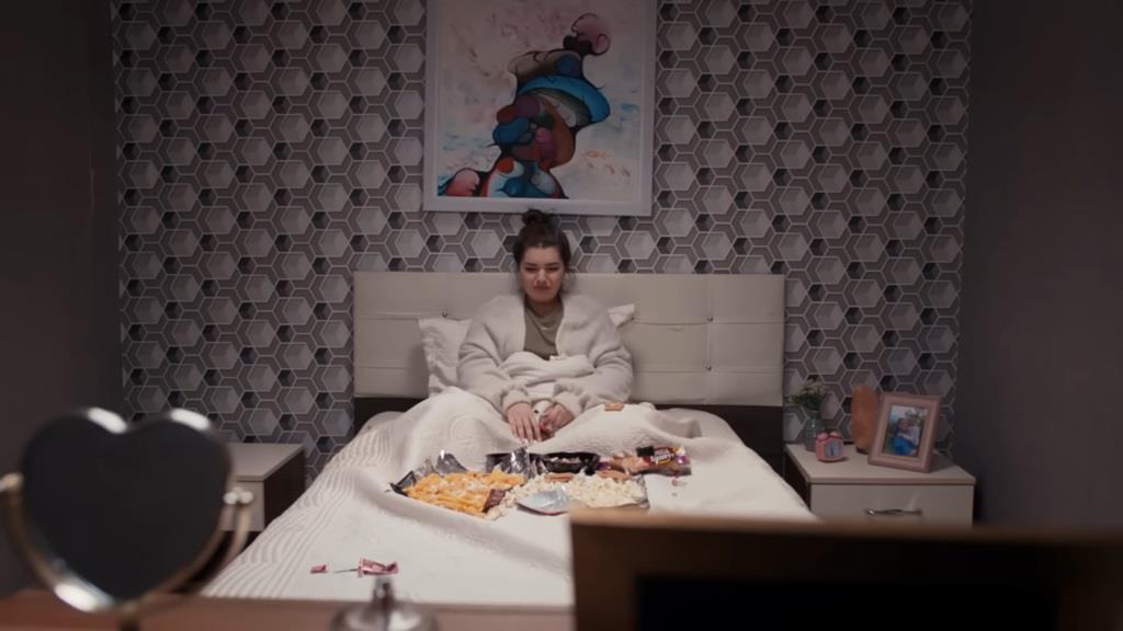 Nese siedzi w łóżku, otoczona otwartymi paczkami pełnymi słodyczy i przekąsek, które kuszą kolorowymi opakowaniami.