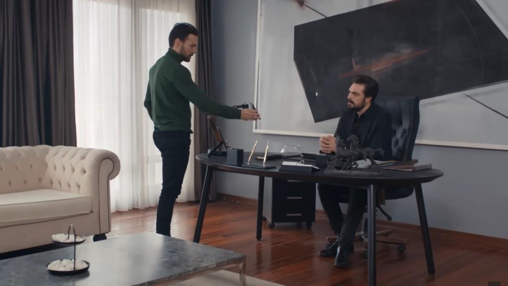 Kubilay stoi przed biurkiem Yamana i pokazuje mu swój telefon.