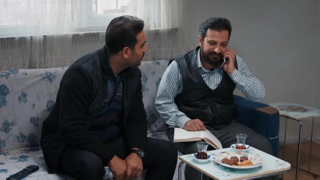 Idris i Kazim siedzą na kanapie. Idris rozmawia przez telefon. Przed nimi znajduje się stolik kawowy z dwiema szklankami herbaty i talerzem z ciastkami.