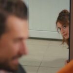 Ege obserwuje Kuzeya, wychylając głowę zza futryny.