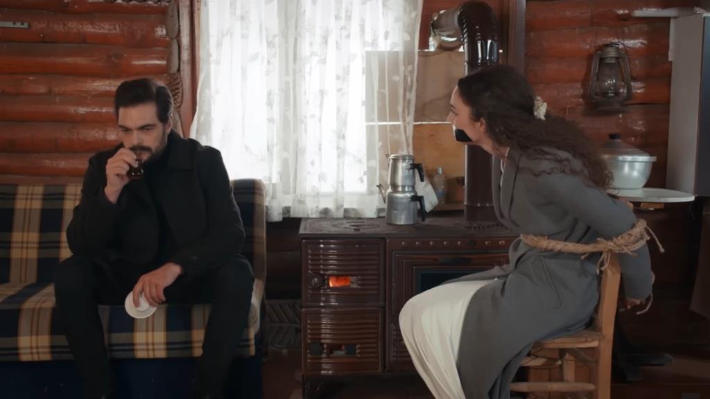 Scena w odosobnionej chacie. Yaman siedzi na wersalce i pije herbatę. Nana jest przywiązana do krzesła.