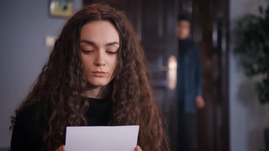 Nana trzyma w rękach wyciągnięte z kosza zdjęcie. W tle widzimy sylwetkę obserwującej ją Akcy.