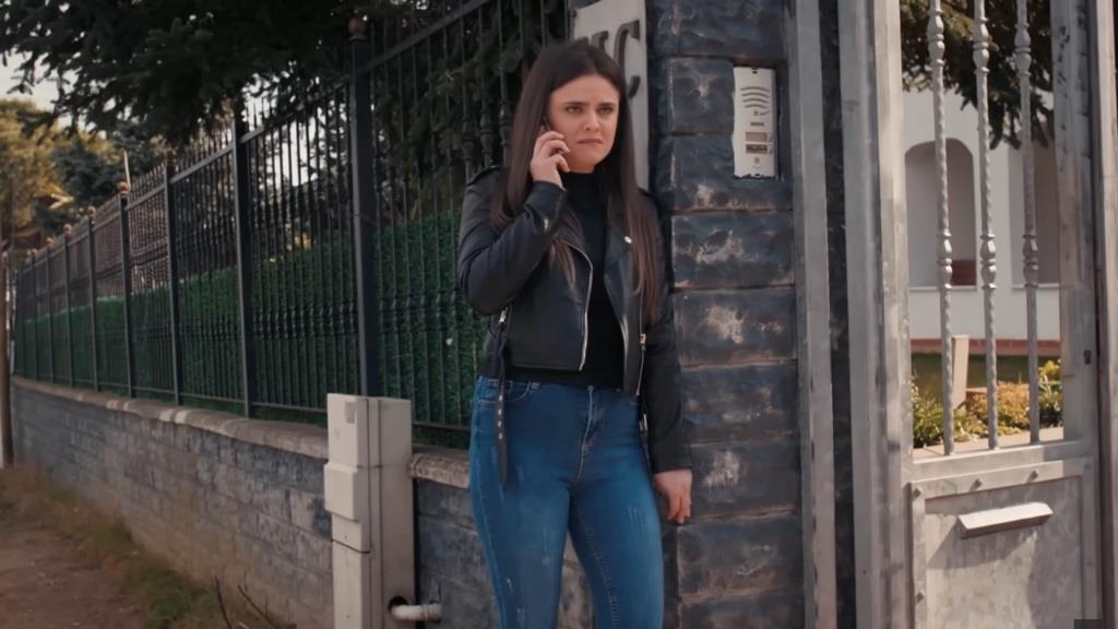 Didem stoi przy ogrodzeniu domu komisarza i rozmawia przez telefon.