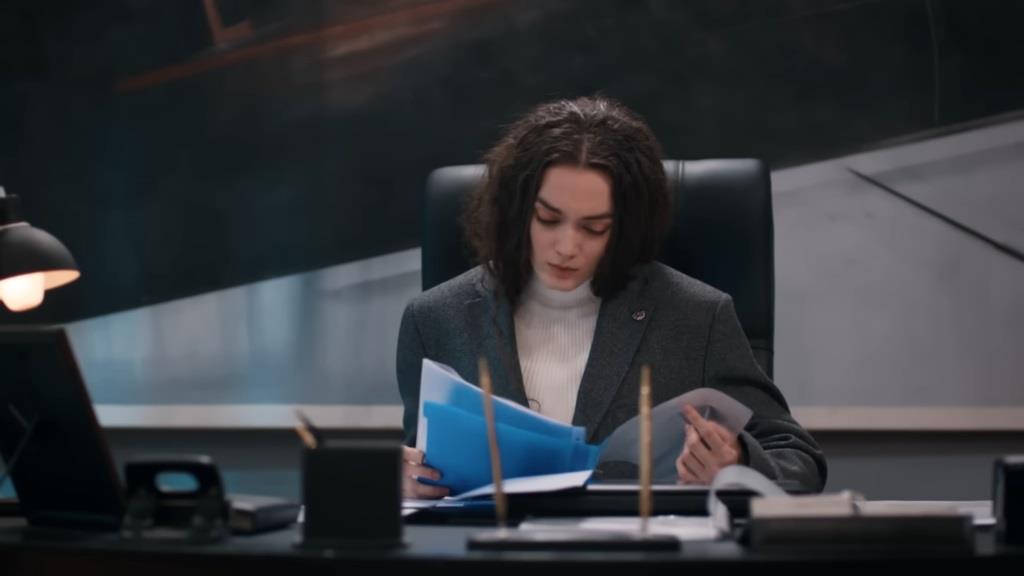 Nana w marynarce Yamana siedzi przy biurku i przegląda dokumenty.