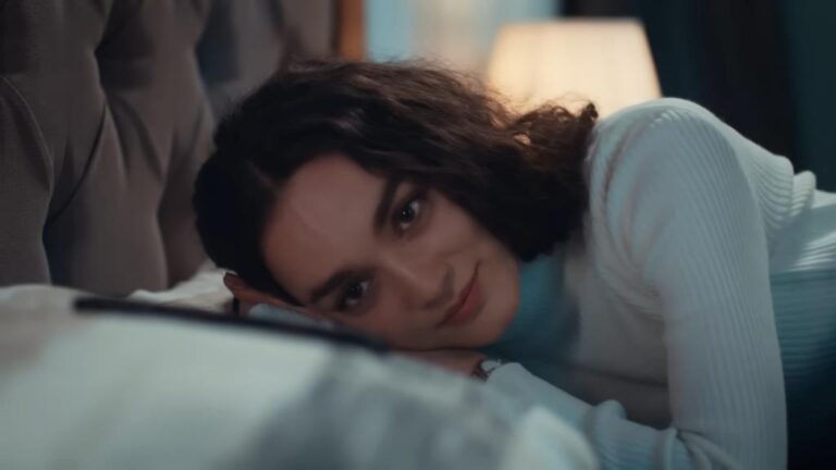 Nana przytula głowę do poduszki i patrzy na położony obok telefon.