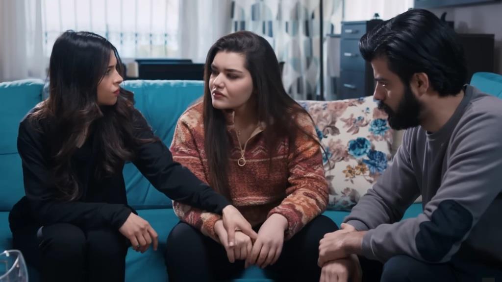 Ayse, Nese i Volkan siedzą na błękitnej kanapie. Ayse trzyma siostrę za rękę.