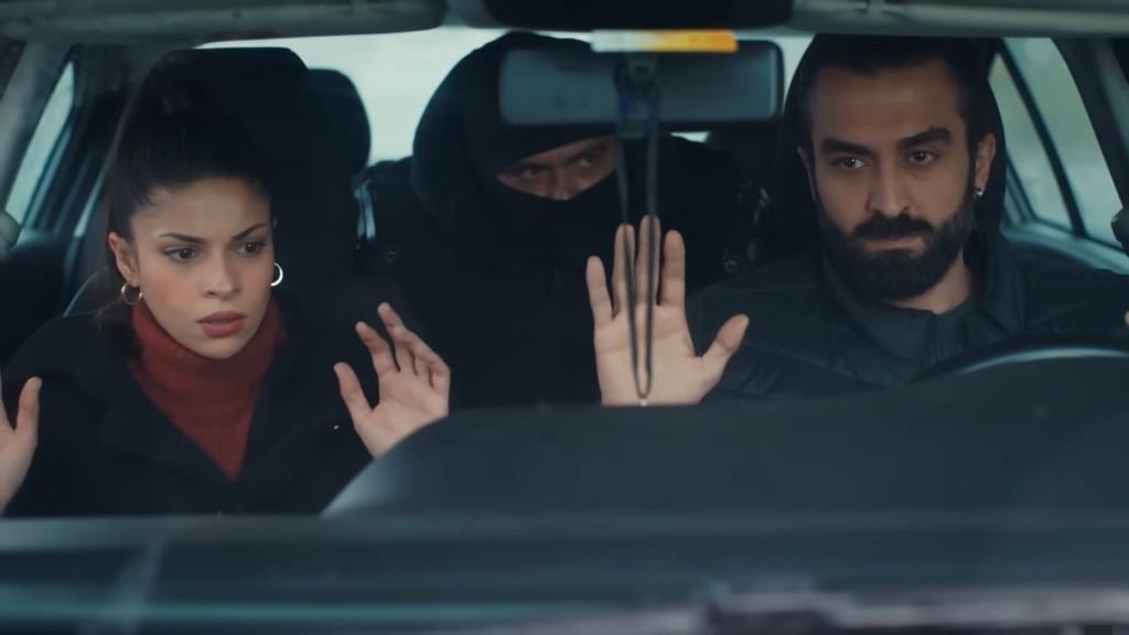 Ayse i Ferit w opałach. Siedząc na przednim rzędzie siedzeń, unoszą ręce do góry, podczas gdy zza ich fotelów wyłania się zamaskowany mężczyzna.