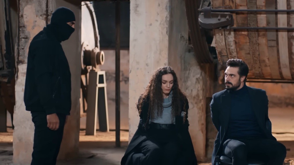 Zamaskowany Kazim stoi obok przywiązanych do krzeseł Yamana i Nany.