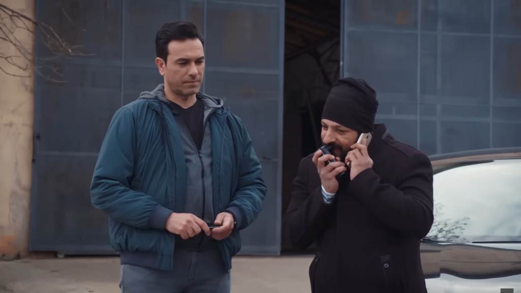 Idris rozmawia przez telefon, używając modulatora głosu. Obok stoi Nedim.