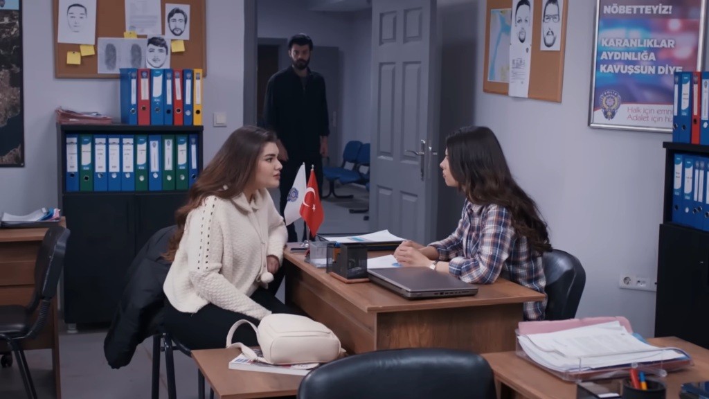 Volkan stoi w wejściu do biura, gdzie przy biurku rozmawiają Ayse i Nese.