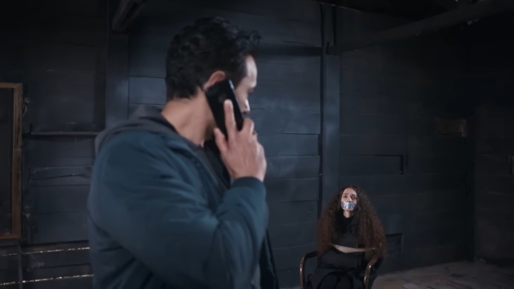 Nedim trzyma telefon przy uchu, patrząc na przywiązaną do krzesła i zakneblowaną Nanę.