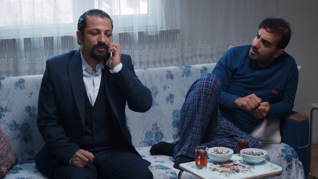 Idris rozmawia przez telefon. Obok niego siedzi Kazim.