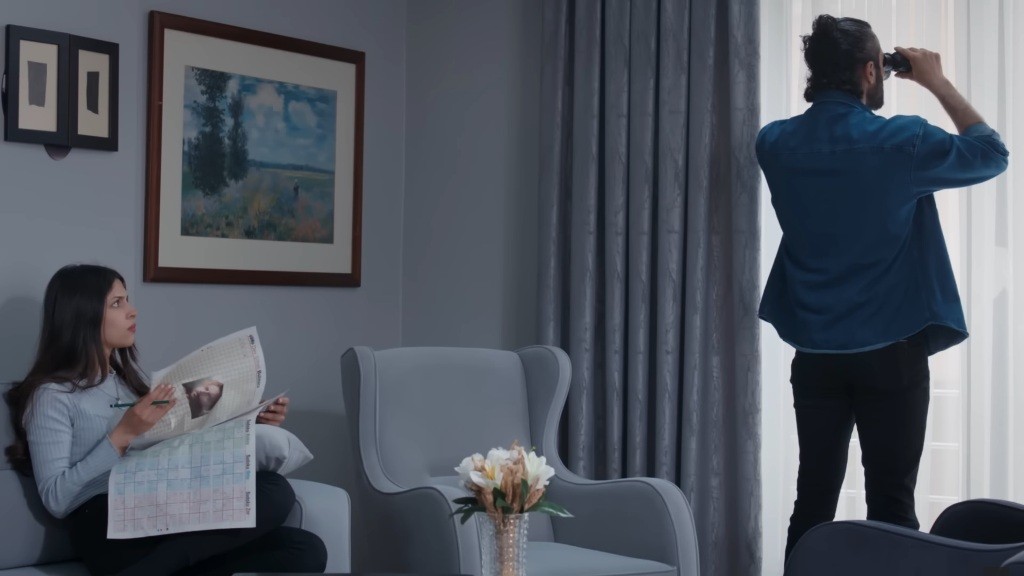 Ferit stoi przy oknie, prowadząc obserwację, a Ayse trzyma w rękach gazetę i patrzy na niego.