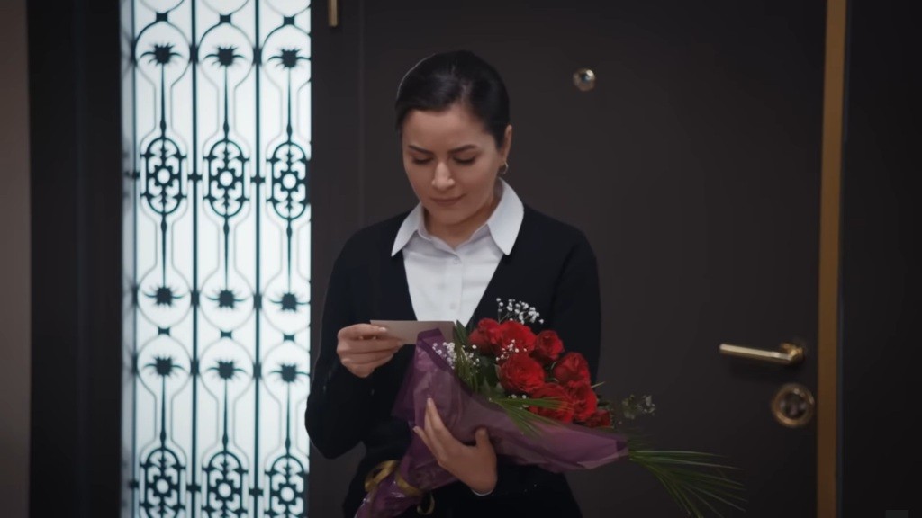 Aynur odbiera bukiet czerwonych róż i czyta dołączony do nich liścik.