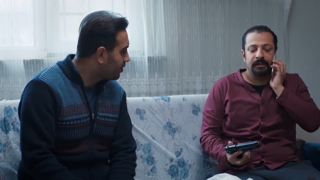 Idris rozmawia przez telefon, trzymając w ręku pistolet. Obok niego siedzi Kazim.