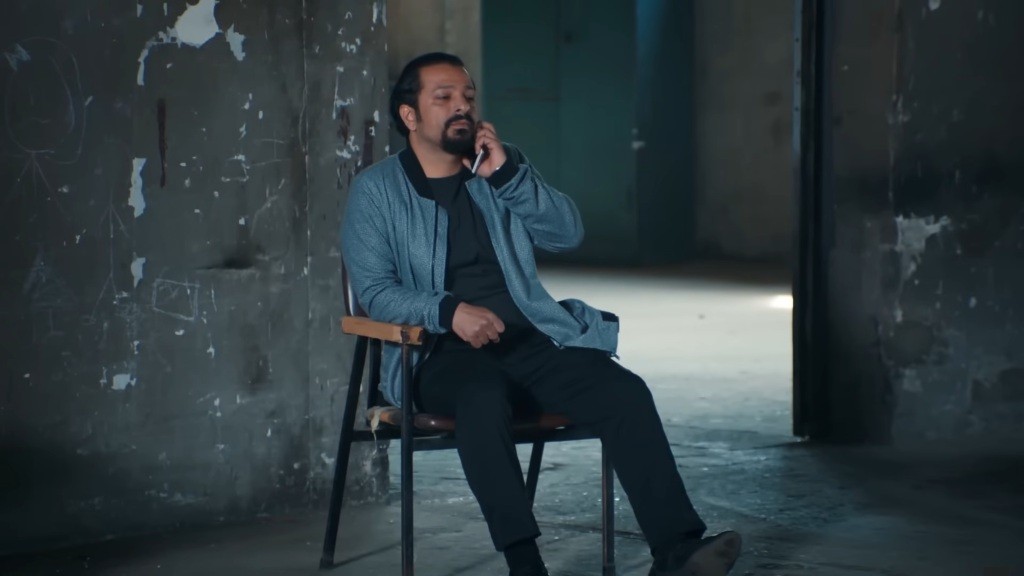 Idris siedzi na krześle w magazynie i rozmawia przez telefon.