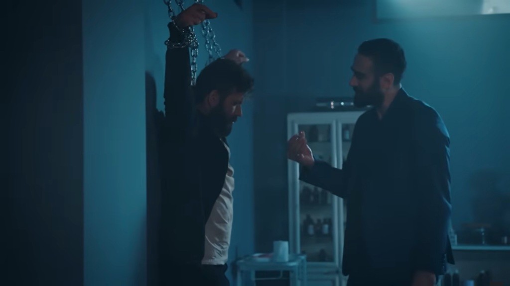 Przykuty do ściany Tunca i stojący przed nim Trucizna.