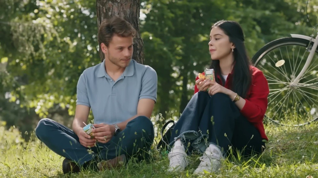 Merve i Cemil siedzą na trawie. Oboje piją soczki.