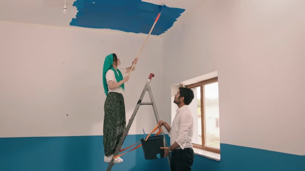 Zeynep stoi na drabinie, malując sufit. Halil trzyma wiadro.