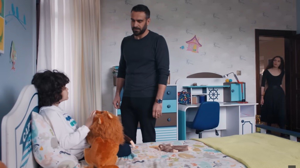 Acar stoi przy łóżku Yusufa, rozmawiając z nim. Przy otwartych drzwiach stoi Nana.