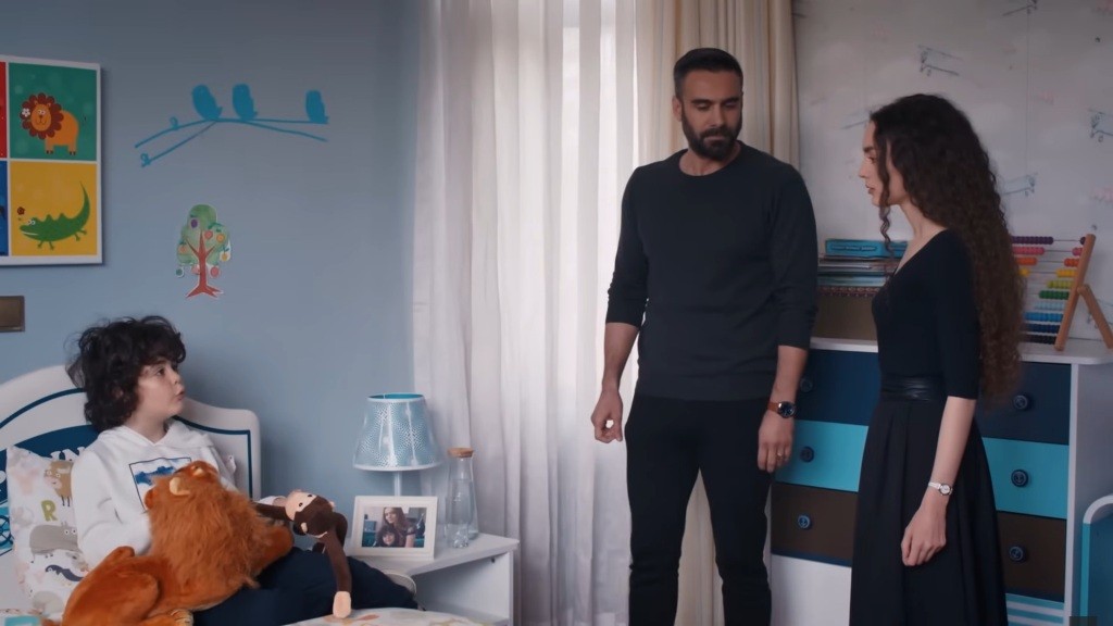 Yusuf siedzi na łóżku, trzymając w rękach pluszowego lwa i małpę. Obok łóżka stoją Acar i Nana.