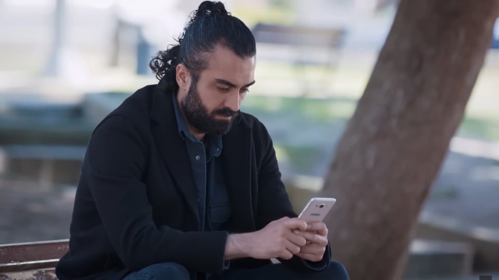 Ferit siedzi na ławce w parku, wpatrując się w ekran telefonu.
