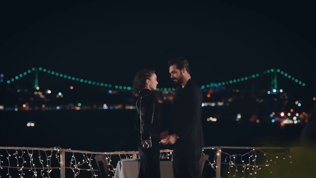 Nana i Yaman stoją naprzeciwko siebie w restauracji i trzymają się za ręce. W tle widać oświetlony Most Bosforski.