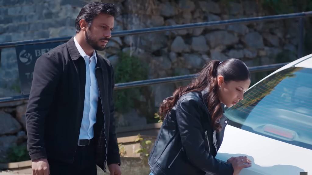 Ayse opiera się o klapę bagażnika samochodu. Za nią stoi Koray.