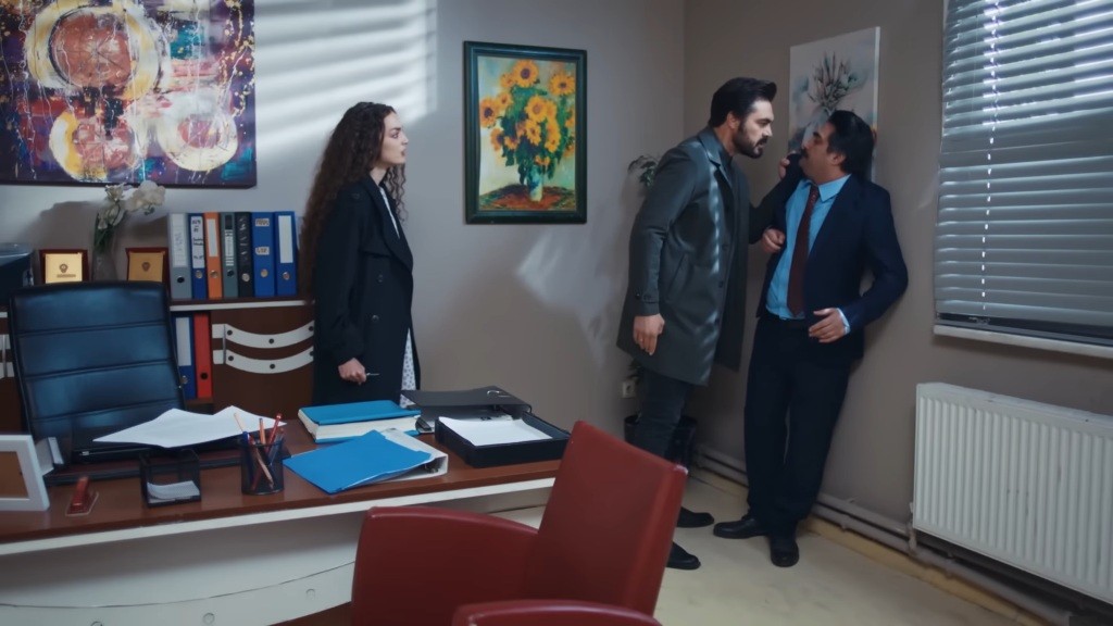 Yaman przyciska grafologa do ściany. Obok stoi Nana.