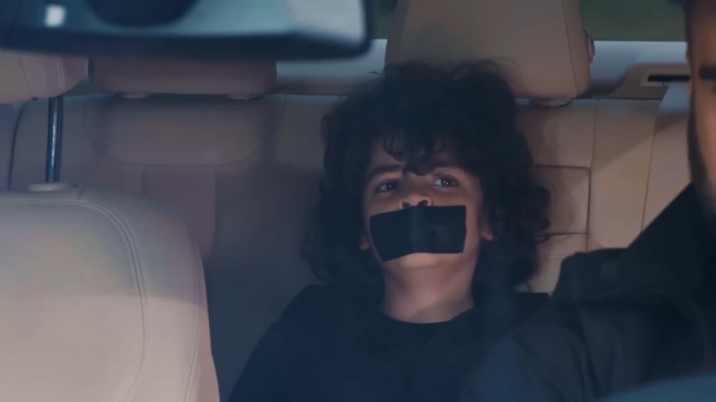 Zbliżenie na Yusufa, siedzącego na tylnej kanapie auta. Usta zaklejone ma taśmą.