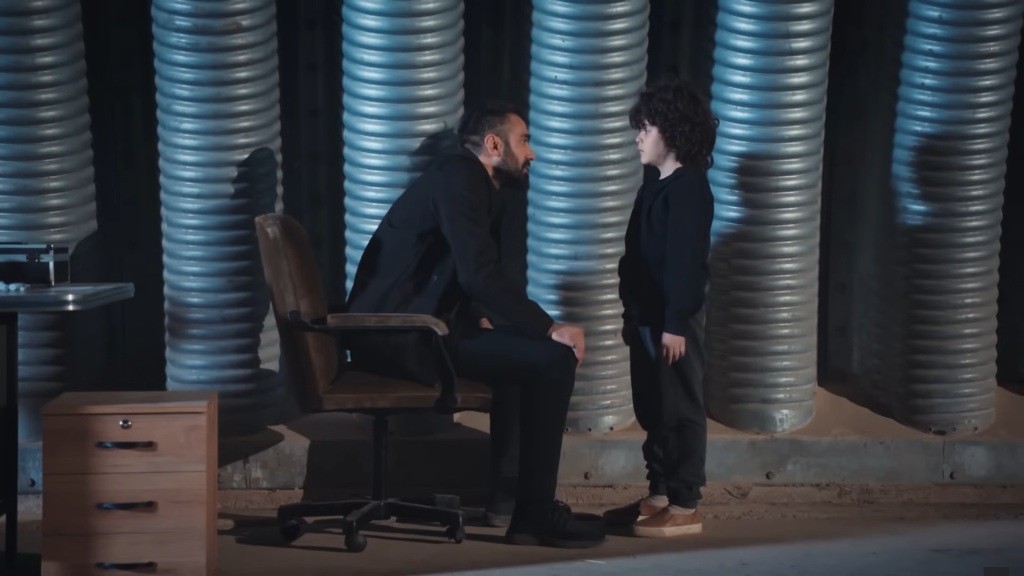 Scena w magazynie. Trucizna siedzi w fotelu, a Yusuf stoi przed nim.