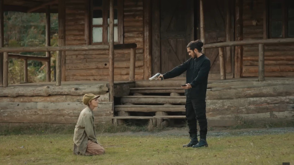 Wilk stoi przed klęczącą na ziemi Naną. W jednym ręku trzyma pistolet, w drugim telefon.