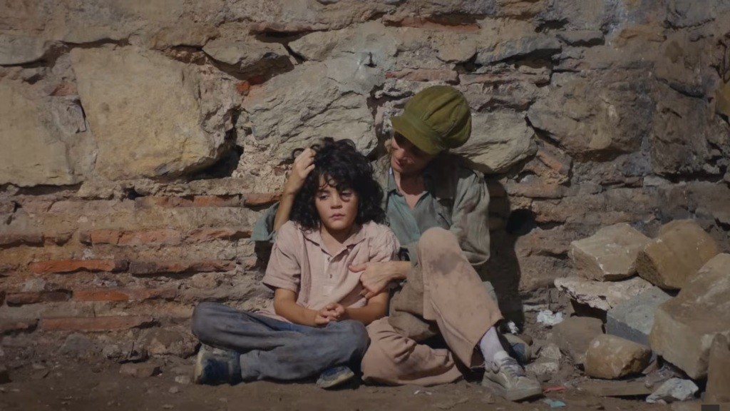 Nana obejmuje Yusufa. Oboje siedzą na ziemi, oparci o kamienny mur.