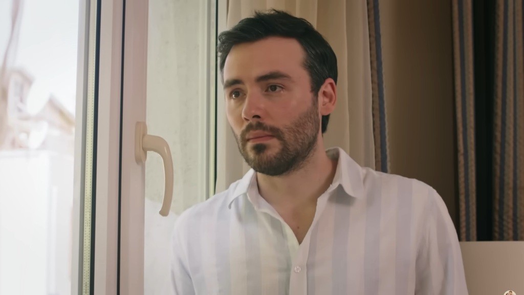 Hakan stoi przy oknie w swoim domu. Wydaje się zakłopotany.
