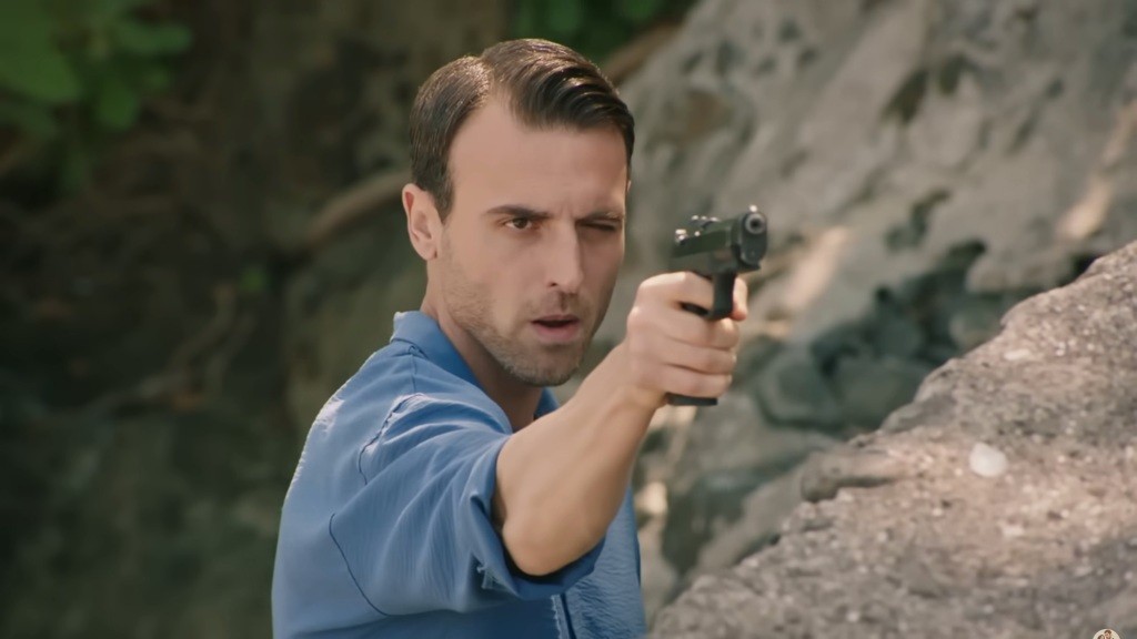 Alper trzyma pistolet w ręku. Przymyka jedno oko, celując.