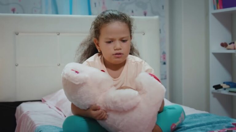 Doga siedzi na łóżku, trzymając pluszaka w rękach.